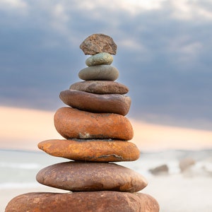 May include: A balanced stack of smooth, reddish-brown stones, resembling a cairn, against a backdrop of a beach and a cloudy sky. The stones vary in size, creating a visually appealing tower.