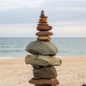 May include: A stone tower built on a sandy beach with the ocean in the background. The tower is made of various sized stones stacked on top of each other.