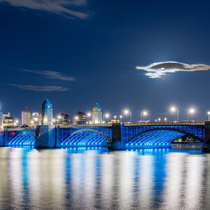 Puede incluir: Una vista nocturna de un puente iluminado en azul con una ciudad en el fondo. La luna es visible en el cielo.