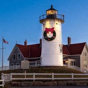May include: A white lighthouse with a red roof and a Christmas wreath on the front. The lighthouse is on a hill with a white picket fence in front of it. There is an American flag flying in the background.