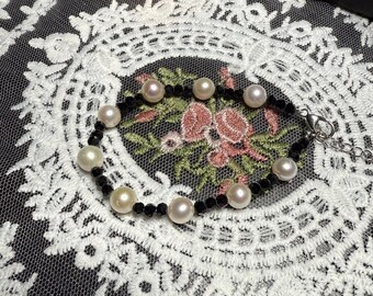 May include: A black and white bracelet with black beads and white pearl accents. The bracelet is on a white lace background.