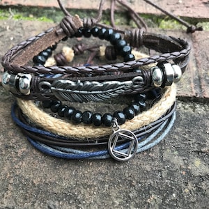 May include: Set of six bracelets featuring a brown leather bracelet with a feather charm, a black beaded bracelet, a brown leather cord bracelet, a beige rope bracelet, a blue cord bracelet, and a silver charm with a square symbol.