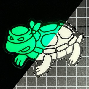 Puede incluir: Una pegatina que brilla en la oscuridad con un diseño de tortuga de dibujos animados. La tortuga tiene una cabeza y un caparazón verdes, con un vientre y extremidades blancos. La pegatina tiene un contorno negro y el texto "@MatchWitsDesign".