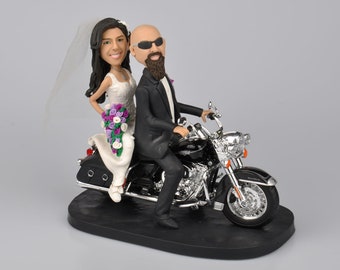 Può includere: Un topper per torta nuziale con una sposa e uno sposo che guidano una motocicletta nera. La sposa indossa un abito bianco e tiene un bouquet di fiori viola e bianchi. Lo sposo indossa un abito nero e occhiali da sole.