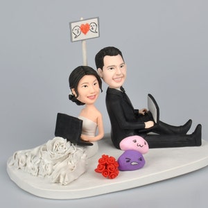 Op de afbeelding: Een bruidstaarttopper met een bruid en bruidegom in een gestileerde cartoon-stijl. De bruid draagt een witte jurk en houdt een laptop vast. De bruidegom draagt een zwart pak en houdt een laptop vast. De topper bevat een bord met een hart en de tekst "Love".