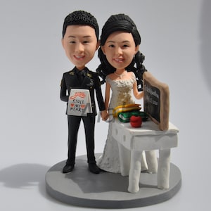 May include: A wedding cake topper featuring a bride and groom in a teacher theme. The bride is wearing a white dress and holding a stack of books. The groom is wearing a military uniform and holding a sign that says "You stole my heart". The cake topper is on a gray base with a small white table and a chalkboard.