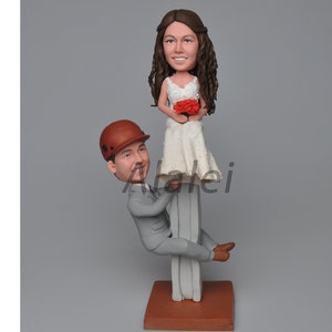 Op de afbeelding: Een bruidstaarttopper met een bruidspaarfiguur. De bruid draagt een witte jurk en houdt een boeket rode rozen vast. De bruidegom draagt een grijs pak en een bruine helm. De figuren staan op een bruine basis.