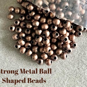 May include: A pile of bronze-colored metal ball-shaped beads with large holes, some spilling from a clear plastic bag. The text "Strong Metal Ball Shaped Beads" is printed in brown at the bottom of the image.