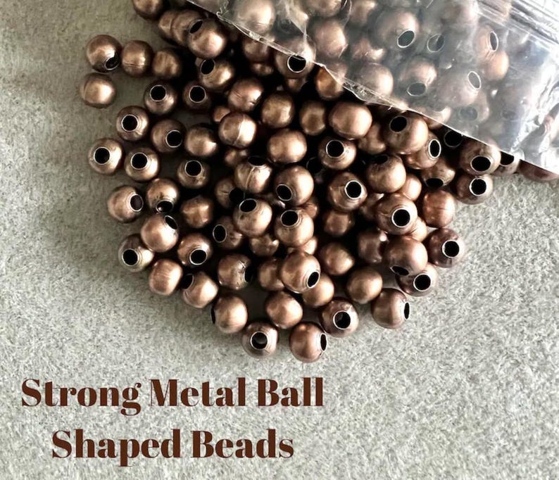 May include: A pile of bronze-colored metal ball-shaped beads with large holes, some spilling from a clear plastic bag. The text "Strong Metal Ball Shaped Beads" is printed in brown at the bottom of the image.