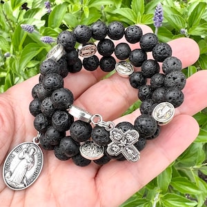 Può includere: Un braccialetto in pietra lavica nera con ciondoli religiosi color argento e un pendente a forma di croce. Il braccialetto presenta perline rotonde e porose, tenuto in mano.
