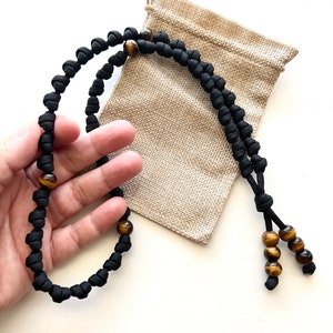 Op de afbeelding: Een zwarte paracord ketting met tijgeroog kralen. De ketting is geknoopt en heeft een kwast aan het einde.