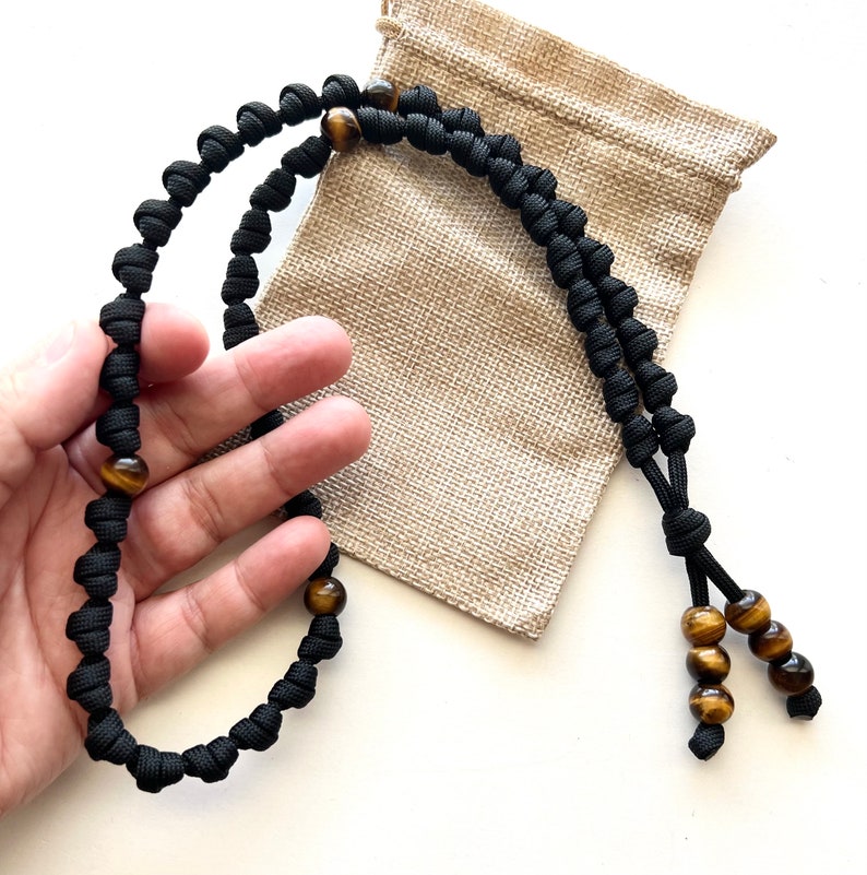 Op de afbeelding: Een zwarte paracord ketting met tijgeroog kralen. De ketting is geknoopt en heeft een kwast aan het einde.