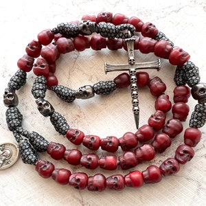 May include: A rosary featuring burgundy skull beads and silver-toned skull accents. A silver cross pendant with skull detailing hangs from the center. A silver medallion with an image is also present. The rosary is displayed on a light-colored surface.