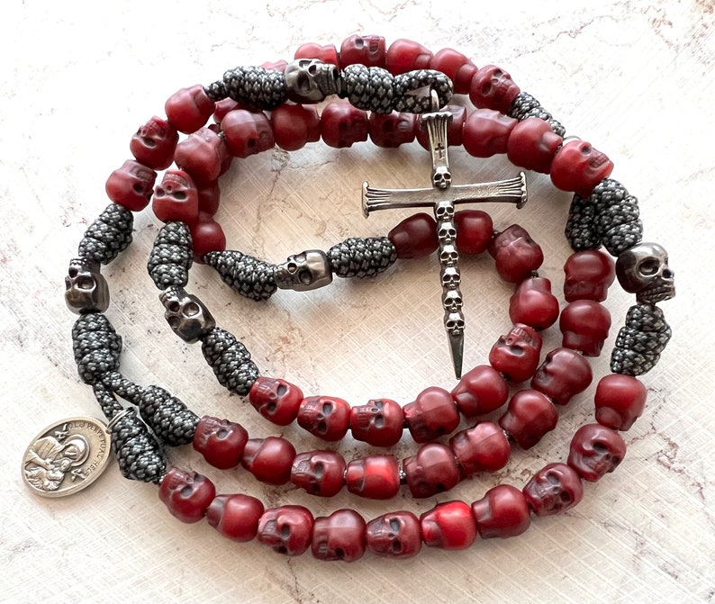 May include: A rosary featuring burgundy skull beads and silver-toned skull accents. A silver cross pendant with skull detailing hangs from the center. A silver medallion with an image is also present. The rosary is displayed on a light-colored surface.
