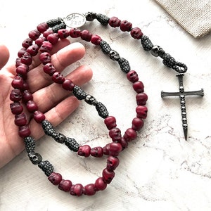May include: A burgundy rosary with skull-shaped beads and a silver cross pendant. The rosary features silver accents and a small metal emblem with the text "Pray for Us". The beads are strung together with a dark cord.