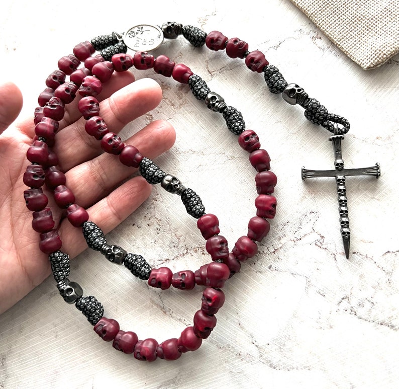 May include: A burgundy rosary with skull-shaped beads and a silver cross pendant. The rosary features silver accents and a small metal emblem with the text "Pray for Us". The beads are strung together with a dark cord.