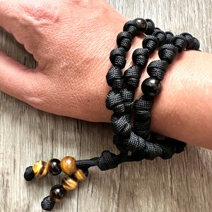 Op de afbeelding: Zwart paracord armband met bruine en gouden tijgeroog kralen. De armband heeft een knoopontwerp en is verstelbaar.