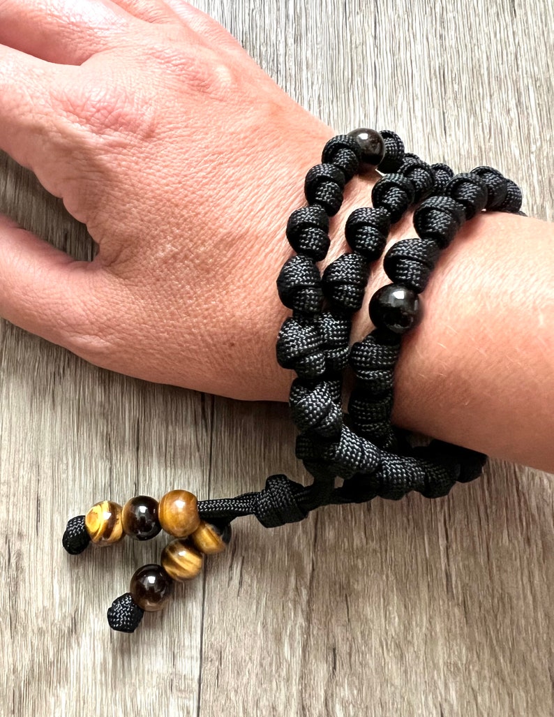 Op de afbeelding: Zwart paracord armband met bruine en gouden tijgeroog kralen. De armband heeft een knoopontwerp en is verstelbaar.