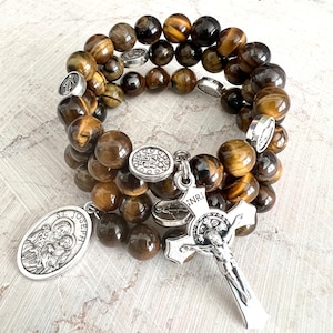 Puede incluir: Un brazalete de triple hebra hecho con cuentas de ojo de tigre y dijes de plata. Los dijes incluyen una medalla de San José, un crucifijo con la inscripción "INRI" y una medalla redonda con una cruz y las letras "S.P.Q.R."