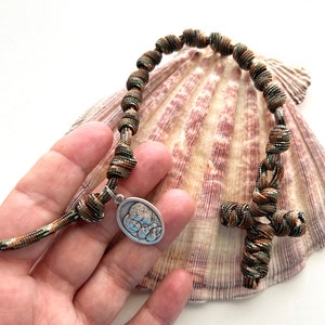 May include: A camouflage paracord rosary with a silver charm and a cross. The rosary is displayed on a seashell.