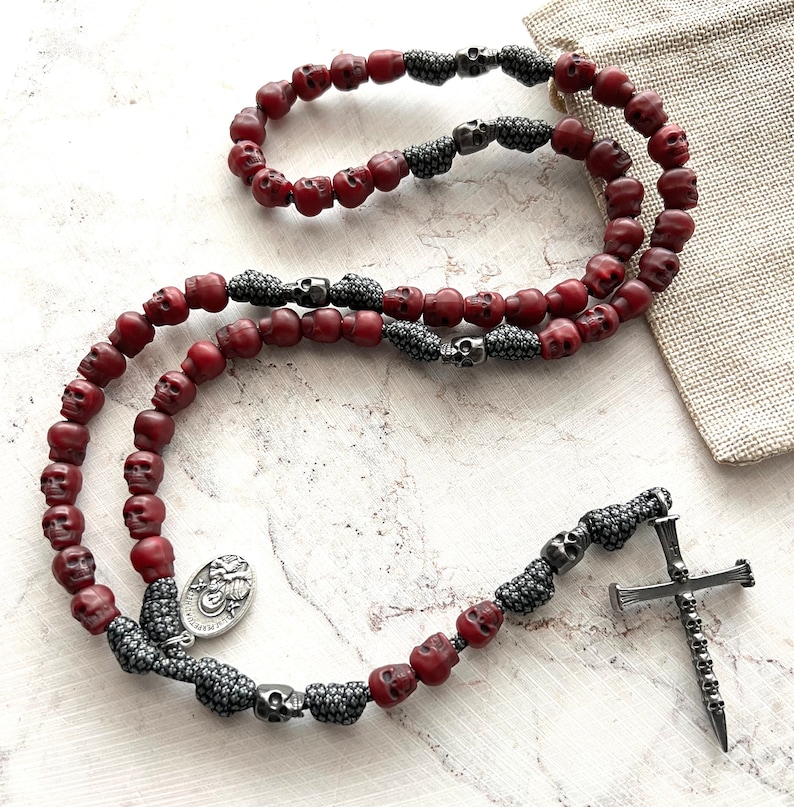 May include: A rosary necklace featuring red skull beads interspersed with silver-toned skull accents. The necklace includes a silver cross pendant with skull detailing and a small religious medallion.