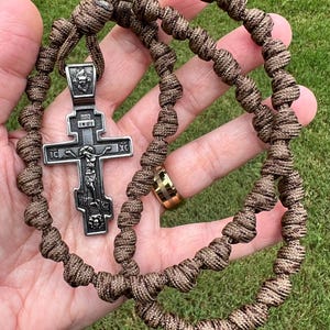Könnte beinhalten: Ein braun-beiges geknotetes Seilrosenkranz mit einem silberfarbenen orthodoxen Kreuzanhänger. Das Kreuz zeigt eine detaillierte Darstellung von Jesus Christus und religiösem Text. Der Rosenkranz wird vor einem grünen, grasbewachsenen Hintergrund gehalten.
