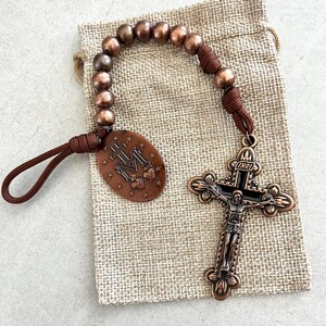 May include: A copper-toned rosary with a detailed crucifix and oval pendant. The pendant features religious symbols and text. The rosary beads are a mix of brown and copper colors. The rosary is resting on a beige burlap bag.