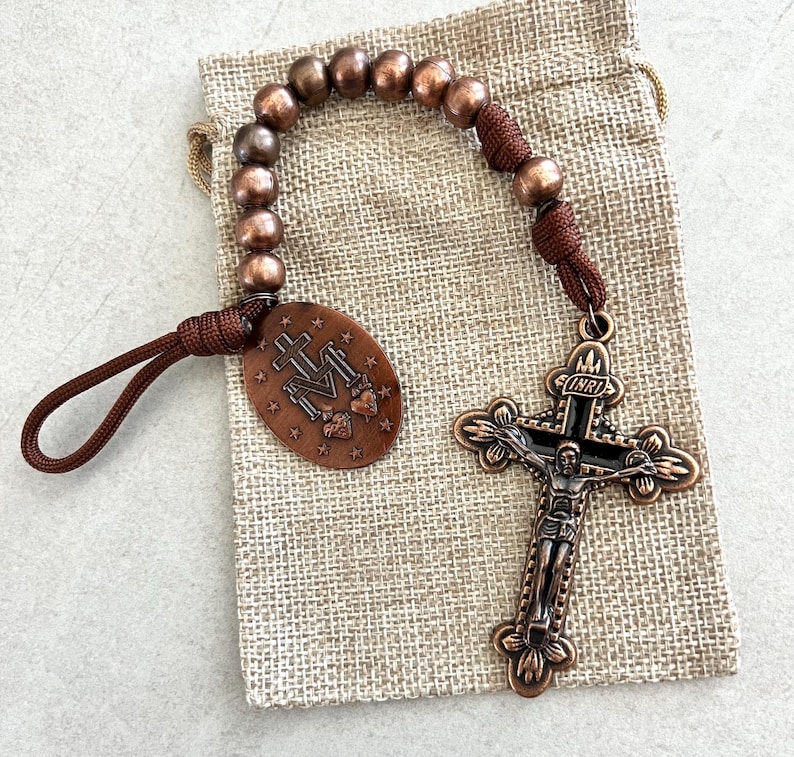 May include: A copper-toned rosary with a detailed crucifix and oval pendant. The pendant features religious symbols and text. The rosary beads are a mix of brown and copper colors. The rosary is resting on a beige burlap bag.