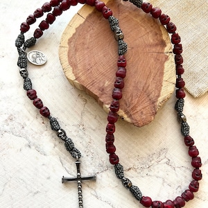 May include: A necklace featuring dark red skull beads and silver-toned accents. The necklace includes a cross pendant and a small circular charm. The beads are strung on a dark cord.