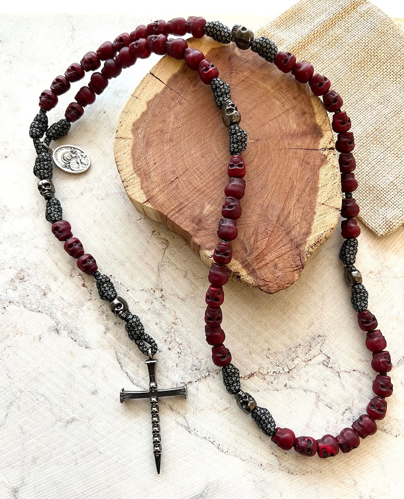 May include: A necklace featuring dark red skull beads and silver-toned accents. The necklace includes a cross pendant and a small circular charm. The beads are strung on a dark cord.