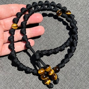 Op de afbeelding: Zwart paracord armband met tijgeroog kralen. Het armband is geknoopt en heeft een kwast aan het einde.