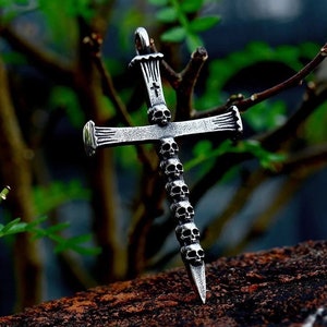 May include: Silver-toned pendant in the shape of a cross, adorned with skull details. The cross features a series of small skulls along its vertical axis and a small cross at the top. The pendant is displayed against a blurred green background.