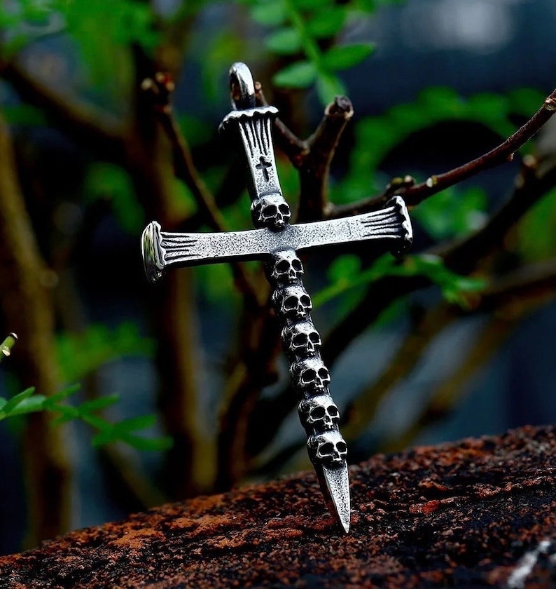 May include: Silver-toned pendant in the shape of a cross, adorned with skull details. The cross features a series of small skulls along its vertical axis and a small cross at the top. The pendant is displayed against a blurred green background.