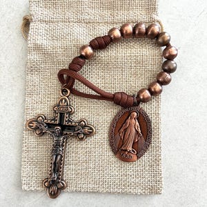 May include: A bronze rosary with a crucifix and a medal featuring a religious figure. The rosary has brown beads and a matching cord. The crucifix has the letters INRI above the figure of Jesus. The rosary is on a small burlap bag.