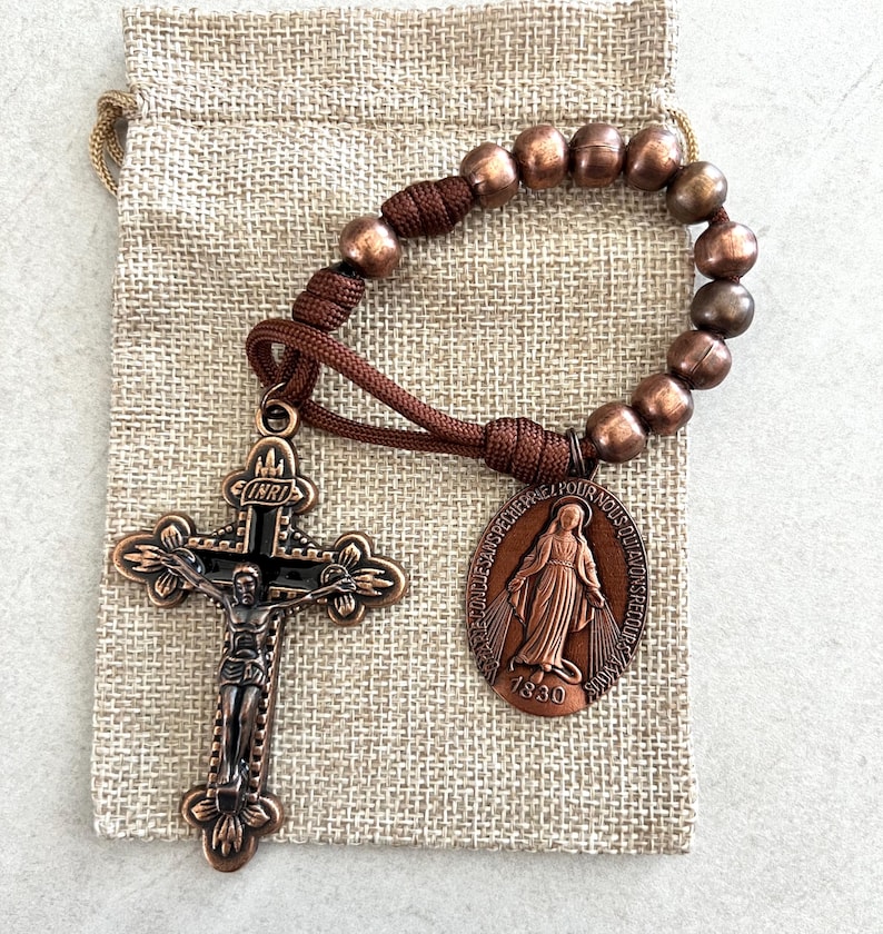 May include: A bronze rosary with a crucifix and a medal featuring a religious figure. The rosary has brown beads and a matching cord. The crucifix has the letters INRI above the figure of Jesus. The rosary is on a small burlap bag.