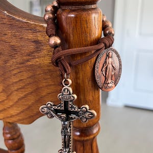 May include: A bronze rosary with a crucifix and a medallion featuring a religious figure. The rosary is wrapped around a wooden post. The crucifix has a detailed depiction of Jesus Christ.