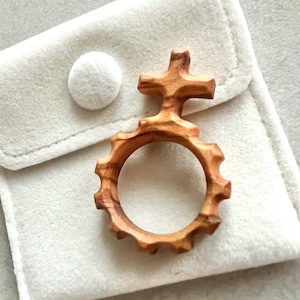 May include: A wooden rosary ring with a cross-shaped top and a circular, gear-like design. The ring is made of light brown wood and rests on a soft, white fabric pouch with a button closure. The rosary ring is a religious item.