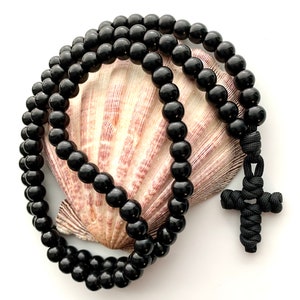 May include: Black prayer beads with a black knotted cross charm. The beads are strung on a cord and are arranged in a circle on a white seashell.