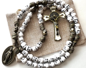 May include: A white skull rosary with a green cord and a metal crucifix with the inscription "INRI". The crucifix features a white figure of Jesus Christ. The rosary is displayed on a brown fabric surface.