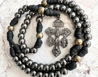 Op de afbeelding: Een zwarte paracord rozenkrans met zilveren metalen kralen en een zilveren kruisbeeld hanger. Het kruisbeeld heeft de inscriptie "JESUS NAZARENUS REX JUDAEORUM" en drie medaillons.