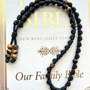 Op de afbeelding: Zwarte gebedskralen met tijgeroogkralen. De kralen zijn geregen aan een zwart koord en hebben een kwast aan het einde. De kralen liggen op een wit oppervlak met de tekst "Our Family Bible" erop gedrukt.