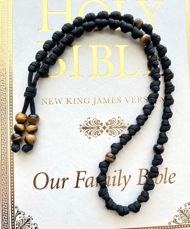 Op de afbeelding: Zwarte gebedskralen met tijgeroogkralen. De kralen zijn geregen aan een zwart koord en hebben een kwast aan het einde. De kralen liggen op een wit oppervlak met de tekst "Our Family Bible" erop gedrukt.