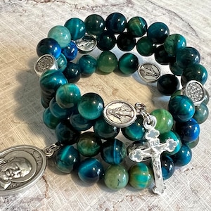 Może przedstawiać: Dwa warstwowe bransoletki wykonane z niebieskich i zielonych koralików z tygrysiego oka z srebrnymi zawieszkami, w tym krzyżem i medalem z napisem "S. PIO" i wizerunkiem twarzy mężczyzny.