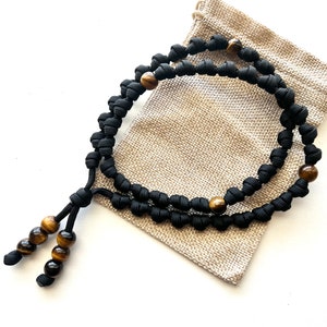 Op de afbeelding: Zwart paracord gebedstouw met tijgeroog kralen. Het touw is geknoopt en heeft een kwast aan het einde. Het touw zit in een kleine jute zak.