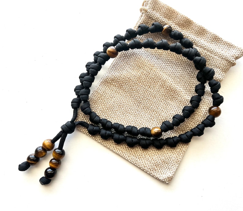 Op de afbeelding: Zwart paracord gebedstouw met tijgeroog kralen. Het touw is geknoopt en heeft een kwast aan het einde. Het touw zit in een kleine jute zak.