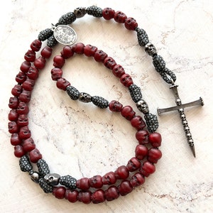 May include: A gothic-style rosary necklace featuring burgundy skull beads, interspersed with silver-toned skull accents and a detailed cross pendant. The necklace includes a silver medallion with an inscription.