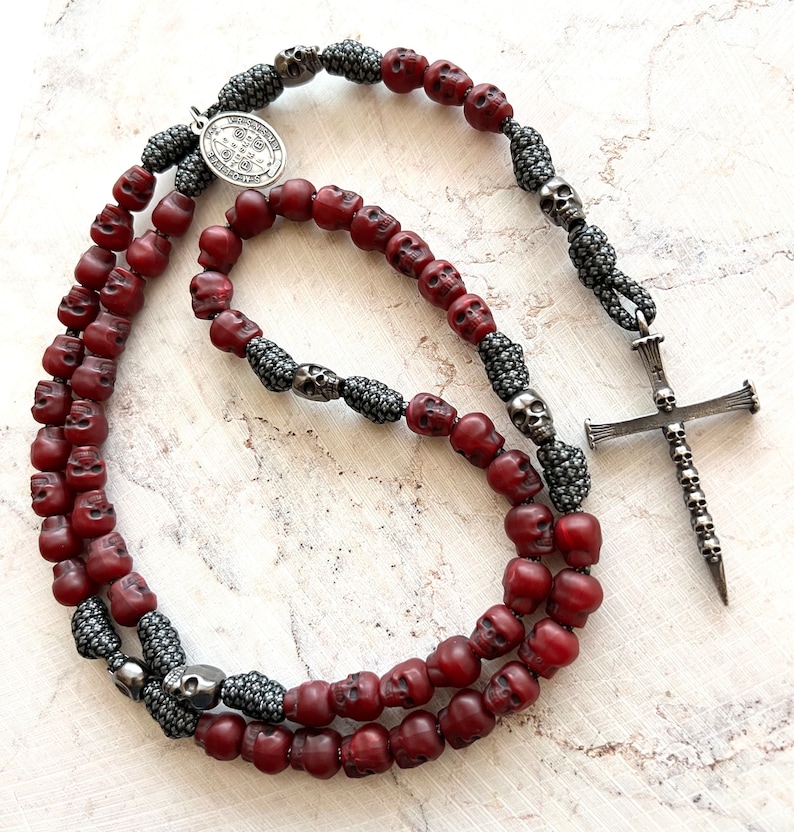 May include: A gothic-style rosary necklace featuring burgundy skull beads, interspersed with silver-toned skull accents and a detailed cross pendant. The necklace includes a silver medallion with an inscription.