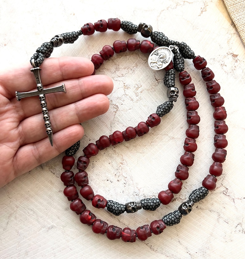 May include: A rosary featuring red skull beads and silver accents. The rosary includes a silver cross pendant with skull detailing and a small circular pendant. The beads are strung on a dark gray cord.