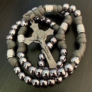 Puede incluir: Un rosario de paracord gris con cuentas plateadas y un crucifijo plateado con la inscripción "INRI".