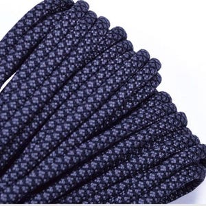 May include: Close-up of a tightly coiled, dark navy blue rope. The rope has a textured surface with a repeating pattern of small, lighter blue squares. The rope is thick and appears durable, suitable for various outdoor and crafting projects.